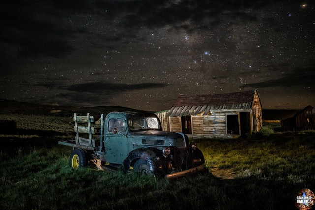 An antique pickup truck sitting in front of a wooden shed. Though a few clouds are overhead the stars are shining brightly.