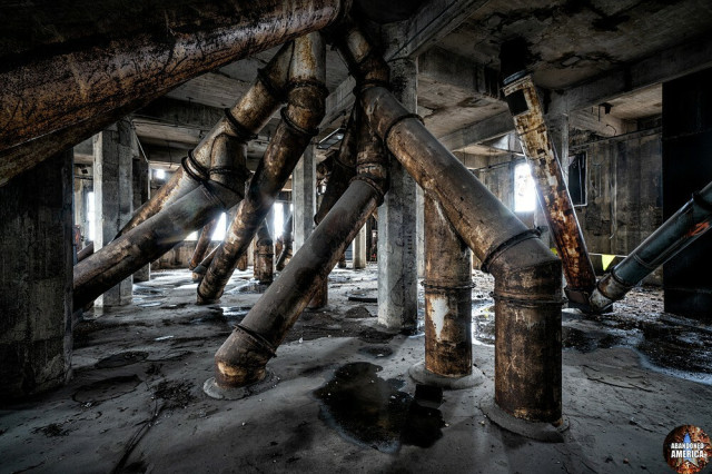 A concrete space with a multitude of enormous rusty branching pipes jutting at odd angles from the ceiling to the floor