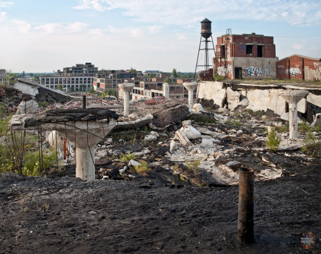 In the foreground, the black tar of the roof of the factory. A few feet away a massive section has collapsed onto the floor below, leaving broken columns and rubble with plants growing out of them. Fluted concrete columns remain standing. In the distance a water tower and more factory buildings are visible.