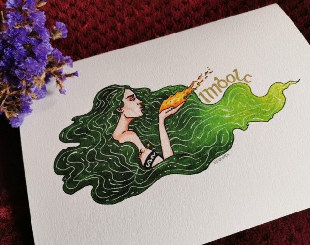 Print of a painting showing a woman with green gradient flowing hair blowing a flame out of her hand, with the word "Imbolc" in gold lettering beside her. The print is on a red velvety background with purple flowers beside it.