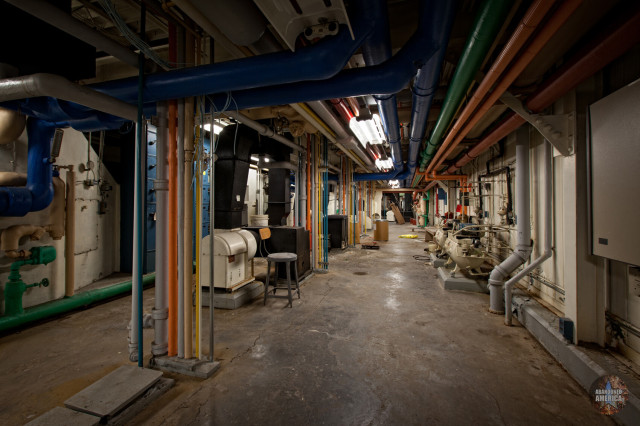 A long hallway with various pipes, wires, fan units, and other bits of large building infrastructure. The most striking aspect are a series of pipes running along the walls and through the floors that are different colors, painted red, blue, yellow, orange, and green