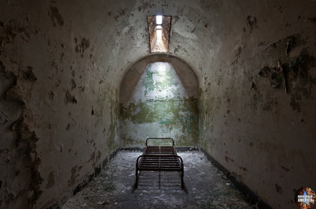 A barrel-roofed cell with a slit in the ceiling creating a shaft of light on a metal bed frame