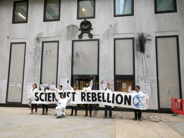 scientists standing outside a building daubed with fake oil holding a sign saying SCIENTIST REBELLION