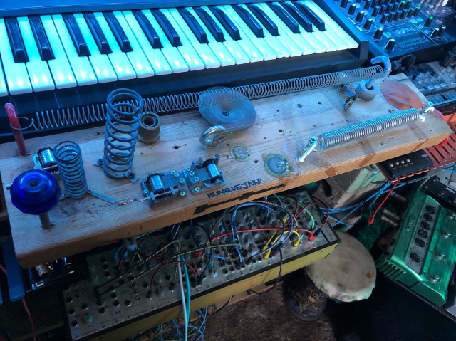 The Hunquejarp: A short 2x6 amplified wooden plank with found objects, springs, and music boxes. It sits in front of a synth keyboard.